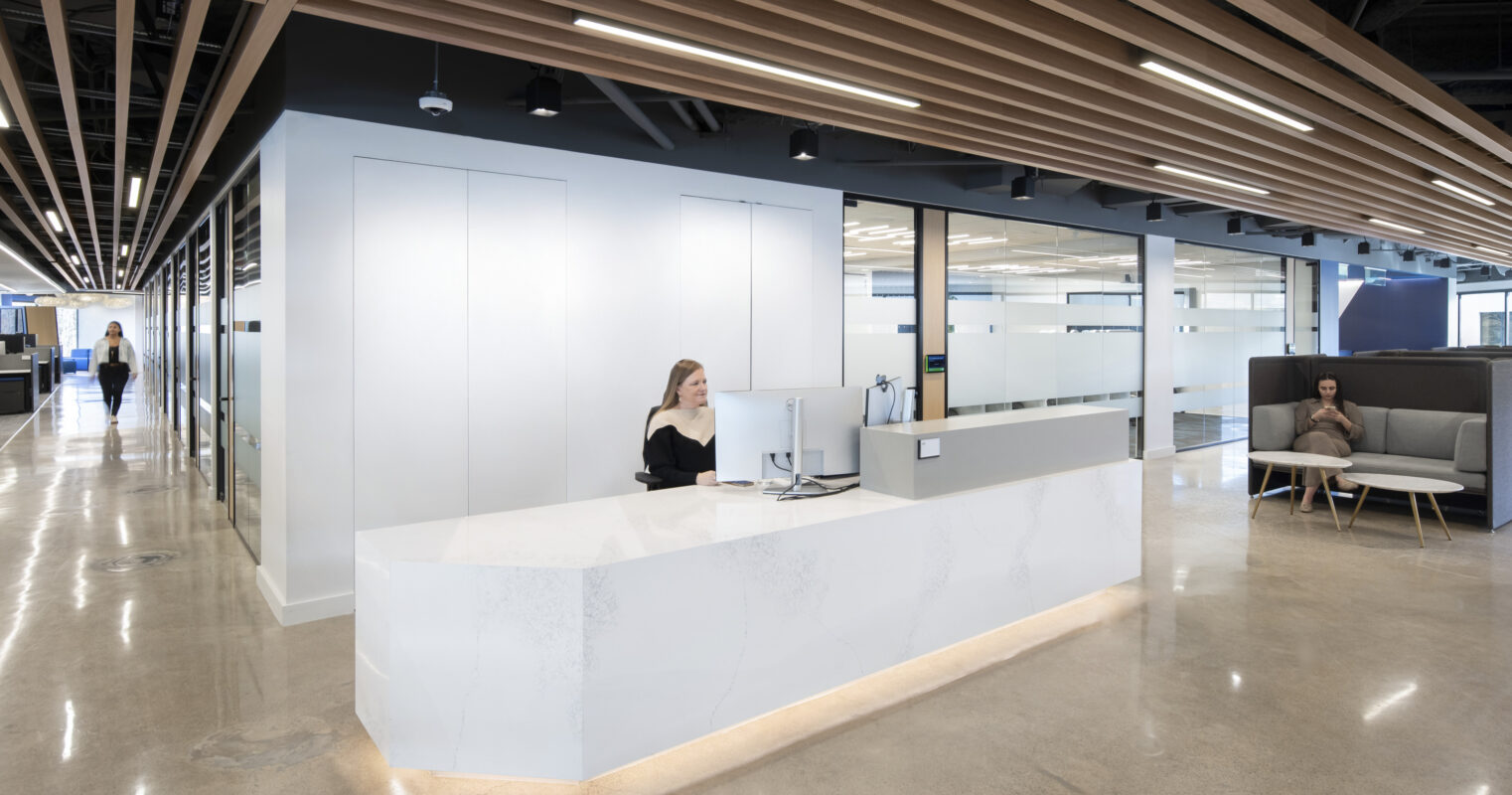 Brightly lit modern office reception area featuring a sleek, angular white reception desk with integrated lighting. The space showcases clean lines, minimalist design, and a neutral color palette, complemented by wooden beams overhead. Comfortable seating and glass partitions create an inviting atmosphere for clients and staff.