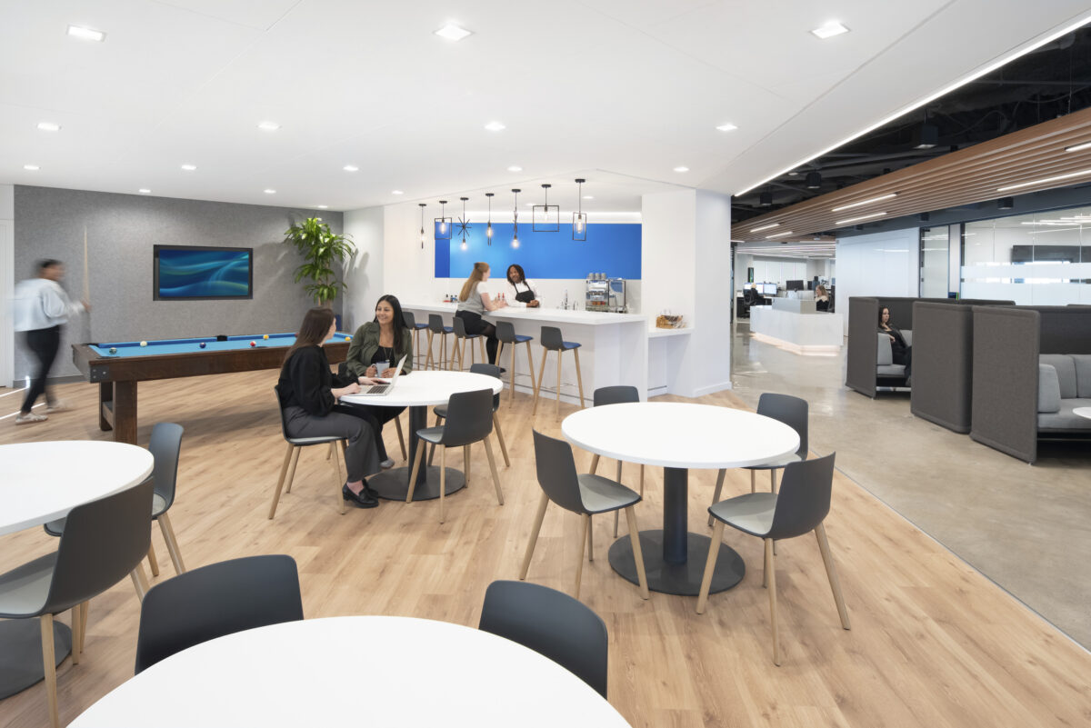 A contemporary office break area features light wood floors, round white tables with modern chairs, and a sleek kitchen bar in the background. Employees engage in conversation at tables while another staff member prepares drinks, creating a vibrant, collaborative atmosphere complemented by a blue accent wall and greenery.