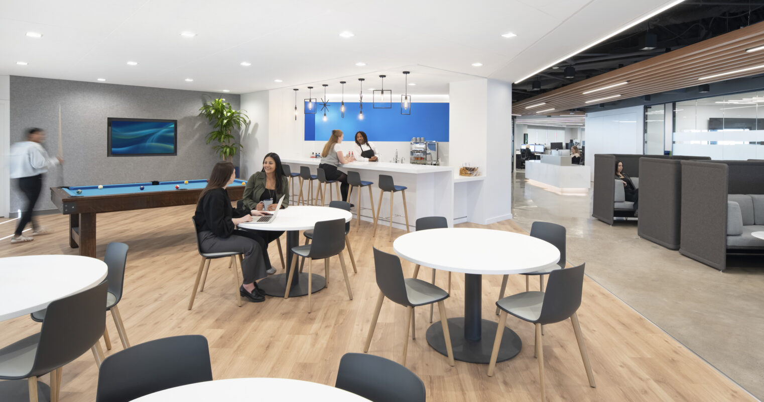 A contemporary office break area features light wood floors, round white tables with modern chairs, and a sleek kitchen bar in the background. Employees engage in conversation at tables while another staff member prepares drinks, creating a vibrant, collaborative atmosphere complemented by a blue accent wall and greenery.