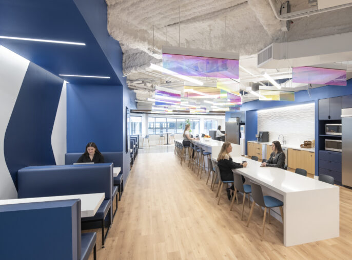 Bright and modern office kitchen and break area featuring blue accent walls and sleek cabinetry. Innovative, multicolored light fixtures suspended from the ceiling illuminate the space, creating a vibrant atmosphere. Collaborative seating areas provide a comfortable setting for informal meetings, enhancing the overall functionality and aesthetic appeal of the design.