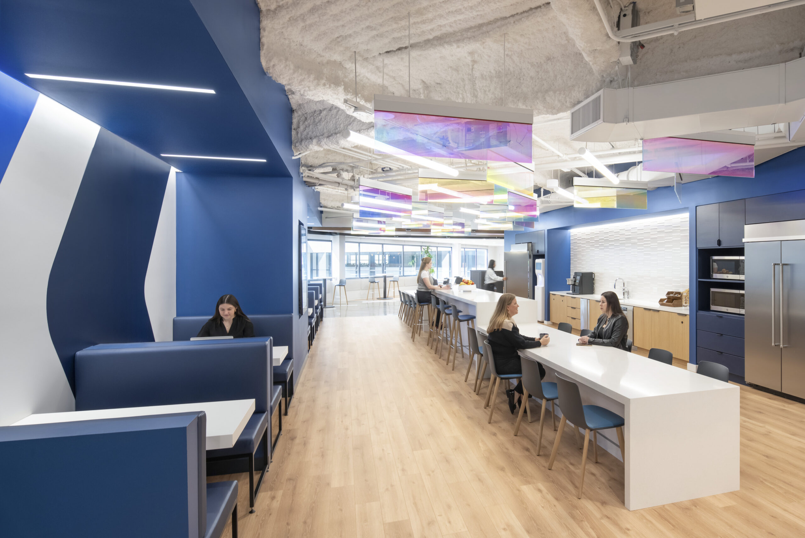 Bright and modern office kitchen and break area featuring blue accent walls and sleek cabinetry. Innovative, multicolored light fixtures suspended from the ceiling illuminate the space, creating a vibrant atmosphere. Collaborative seating areas provide a comfortable setting for informal meetings, enhancing the overall functionality and aesthetic appeal of the design.