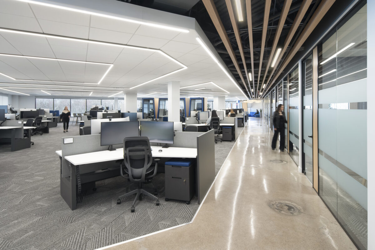Bright and spacious contemporary office interior showcasing an open-plan layout. Workstations feature ergonomic chairs and dual monitors, enhancing functionality. Natural light flows through large windows, complemented by stylish geometric ceiling lights, while polished concrete floors add a modern, industrial feel, promoting a collaborative work environment.
