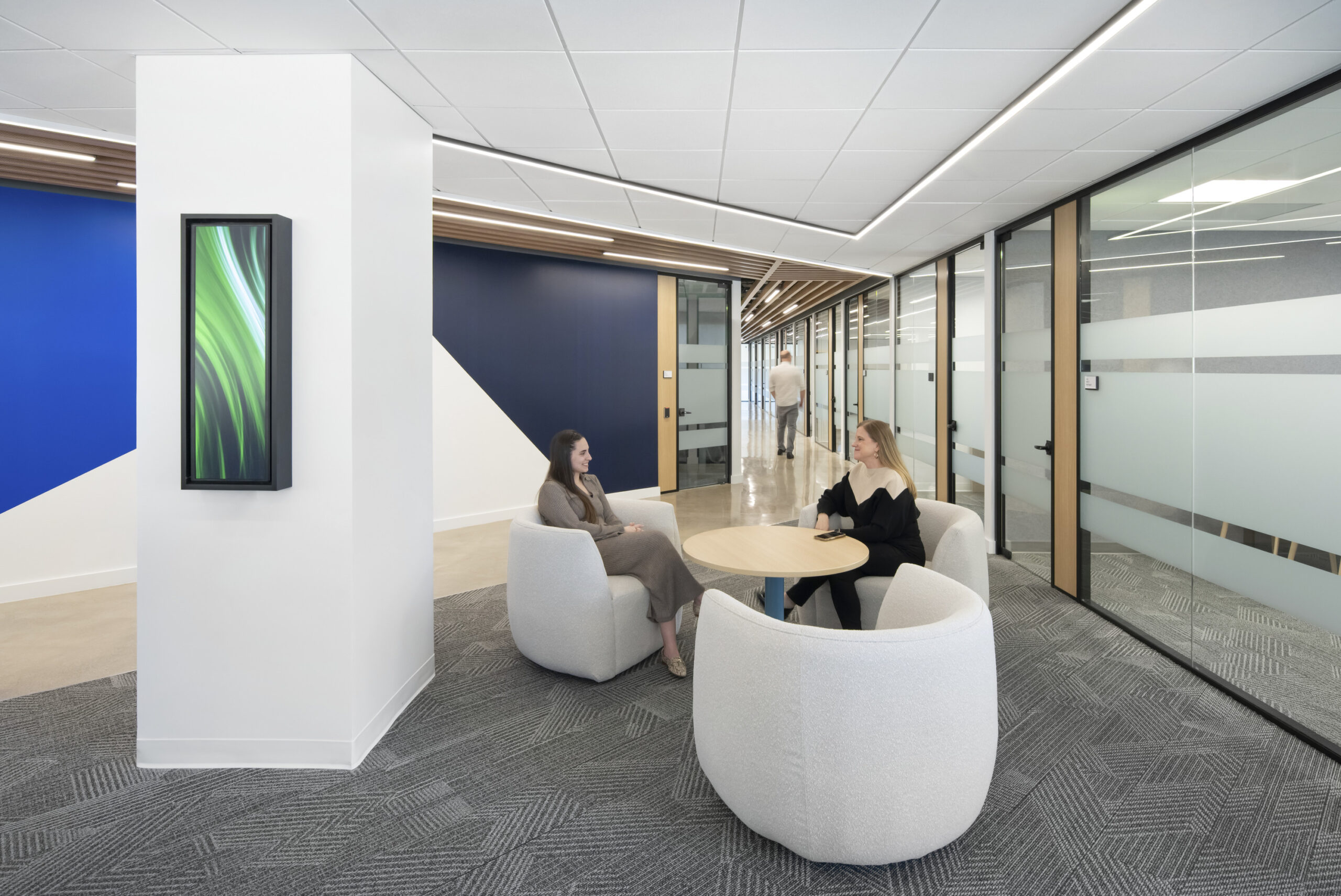 A modern office space featuring two individuals seated at a round wooden table, surrounded by soft, curved white chairs. The backdrop includes a striking blue geometric wall design and sleek glass partitions, enhancing the open concept. Subtle natural lighting complements the contemporary decor, fostering a collaborative atmosphere.