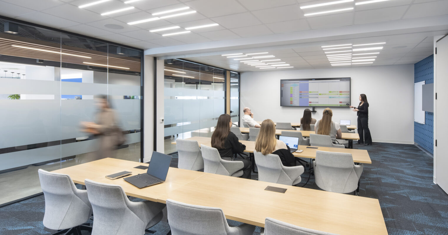 Modern conference room featuring a long wooden table surrounded by ergonomic chairs. Light floods the space through large glass panels, enhancing collaboration. A digital screen displays presentations, while a glass-walled room adds transparency and an open feel, supporting dynamic work environments and fostering teamwork.