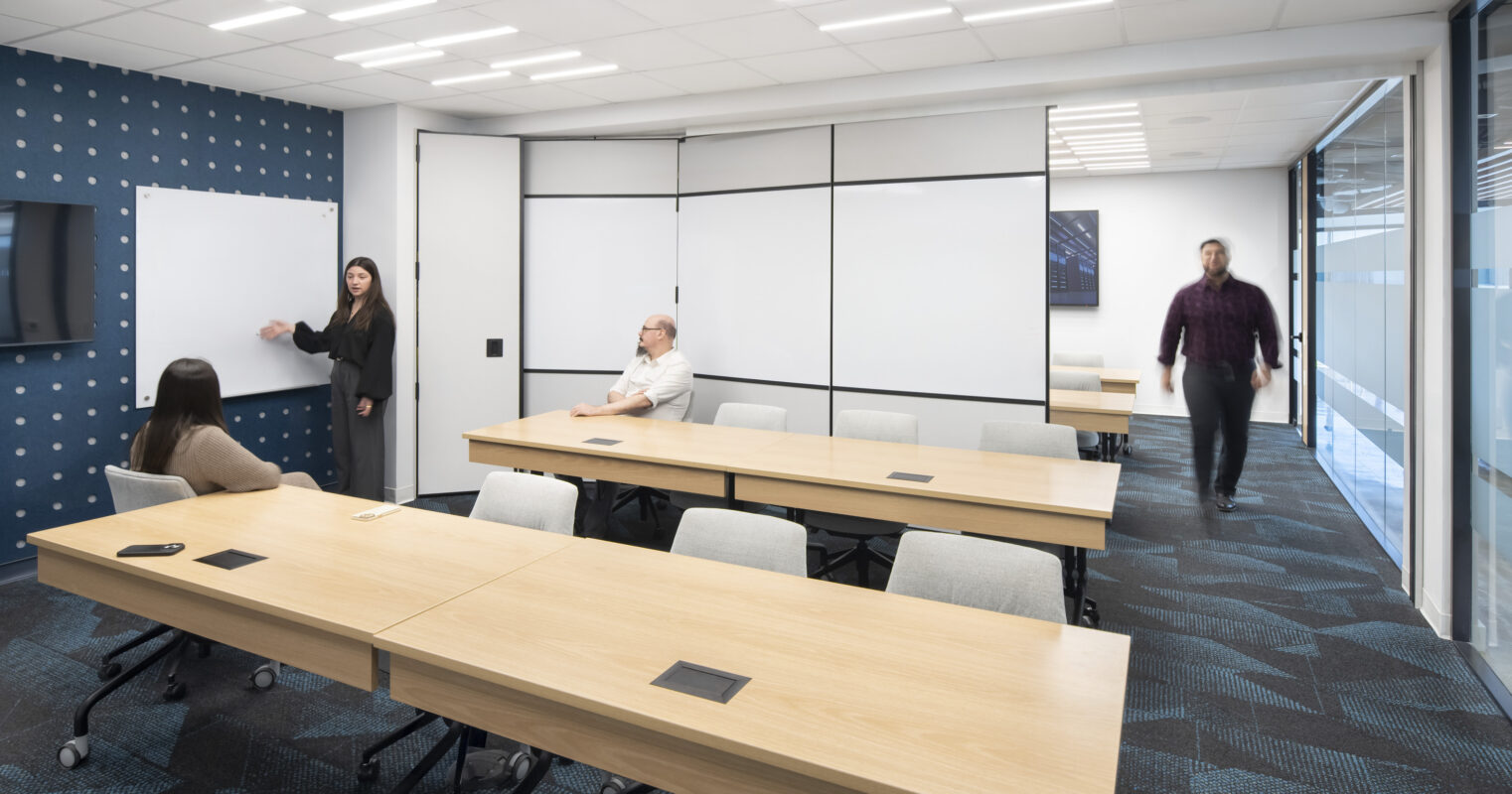 A modern conference room showcasing a collaborative workspace. Features include a series of light wooden tables arranged for group discussions, a dark blue accent wall with a geometric pattern, and large glass windows allowing natural light. Minimalistic design elements and technology-integrated tables enhance functionality and engagement.