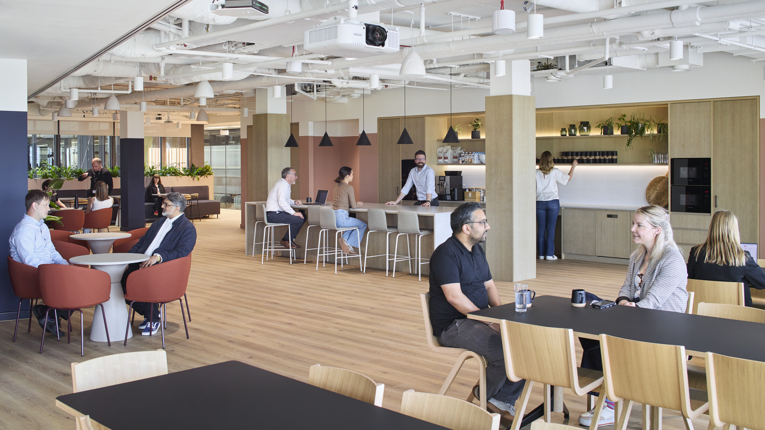 A modern open-plan office showcases a blend of collaborative and private work areas. Warm wood flooring complements sleek furniture in neutral tones, while greenery adds vibrancy. Pendant lighting above a kitchen island highlights a casual dining space, encouraging interaction and informal gatherings among employees. Soft seating areas foster comfort and creativity.