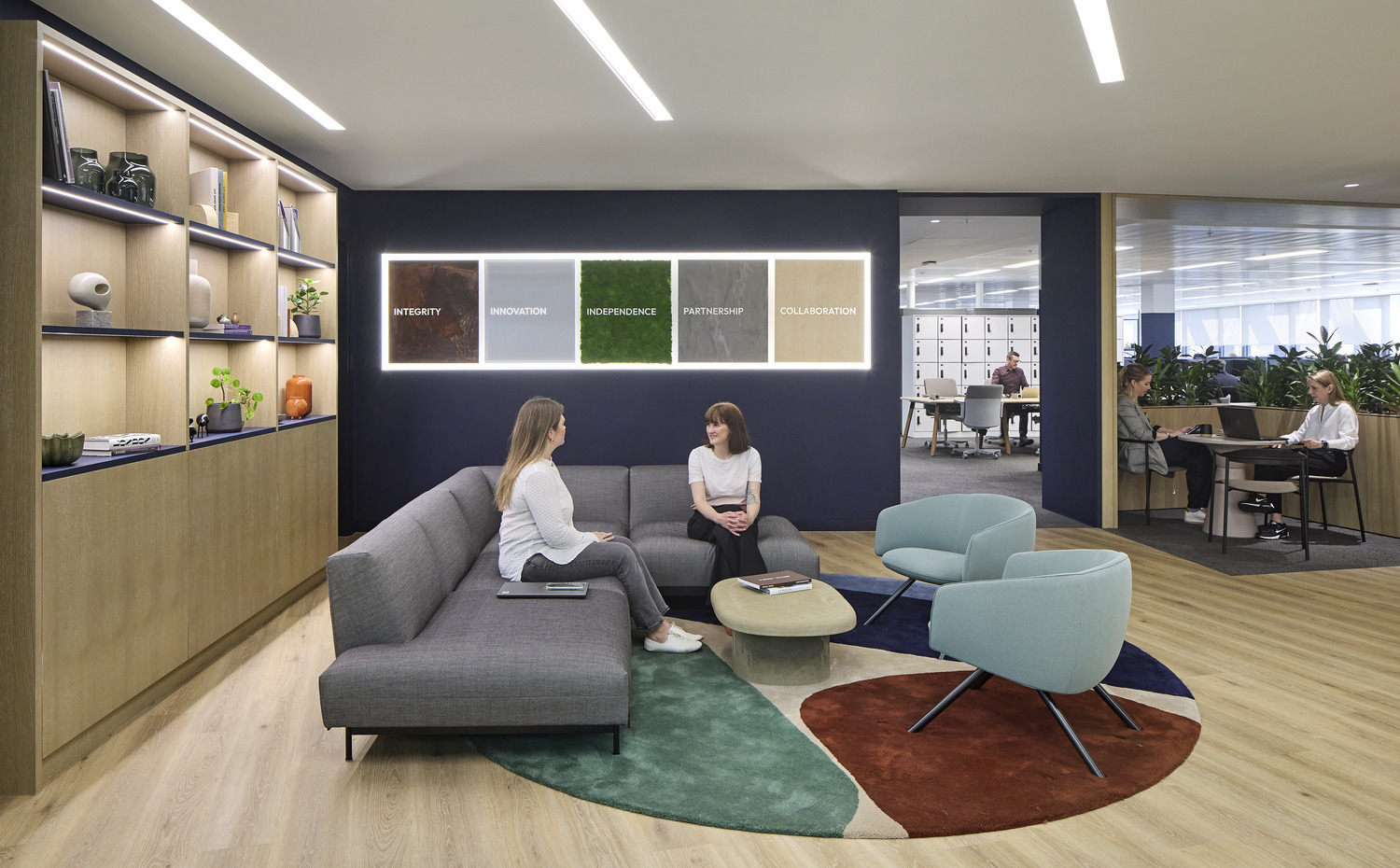 A modern office lounge features a contemporary gray sectional sofa with two people engaged in conversation. Behind them, a wall displays illuminated panels featuring words like "Integrity" and "Innovation." The space includes stylish planters and a colorful circular rug, enhancing a collaborative and inviting atmosphere.