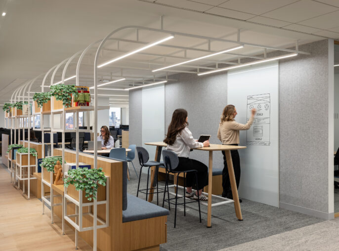 A modern office space featuring collaborative work zones. Two women engage in creative brainstorming at a standing-height table with minimalist design, surrounded by potted greenery. Clear glass partitions and a soft color palette enhance the open feel, while ergonomic seating and organized shelving promote productivity and comfort.