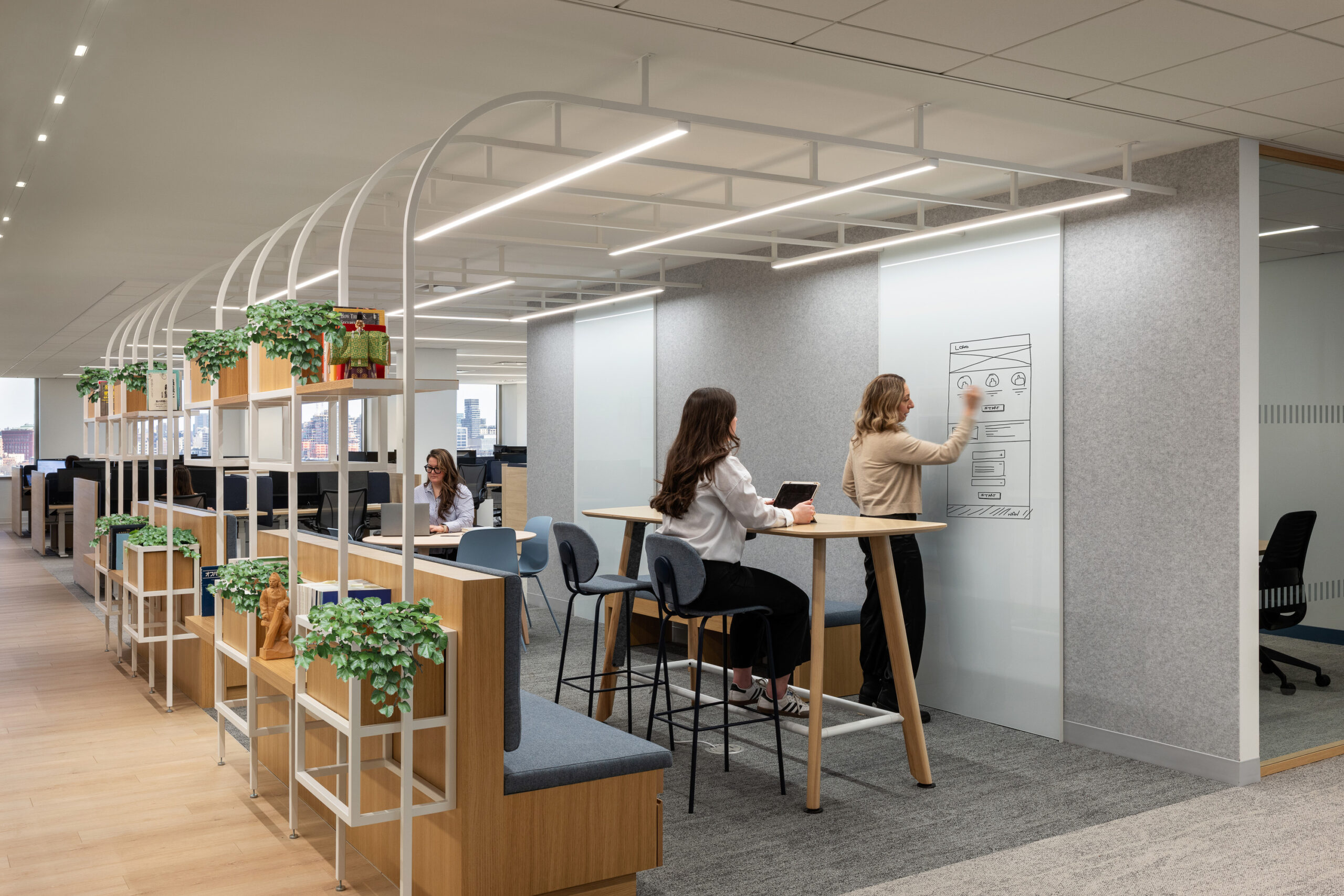 A modern office space featuring collaborative work zones. Two women engage in creative brainstorming at a standing-height table with minimalist design, surrounded by potted greenery. Clear glass partitions and a soft color palette enhance the open feel, while ergonomic seating and organized shelving promote productivity and comfort.