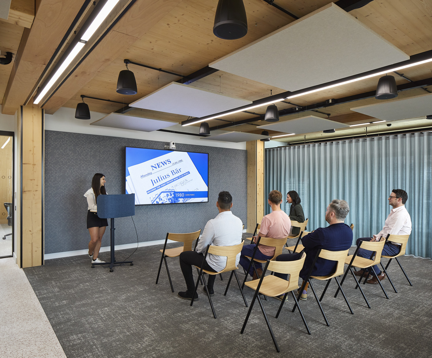 A modern conference room featuring a large digital screen presenting news updates. The space is illuminated by contemporary pendant lights, with wood paneling and acoustic panels enhancing sound quality. Attendees of diverse backgrounds are seated on minimalist wooden chairs, attentively engaged in the presentation.