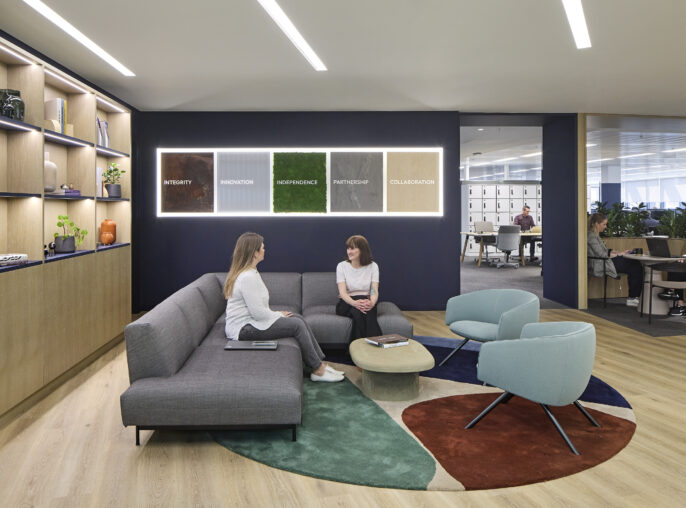 In a modern office lounge, two professionals engage in conversation on a contemporary gray sofa. The space features a stylish circular rug with earth-tone accents and a backdrop of wooden shelves displaying decorative items. Colorful framed words emphasize core values, enhancing the inviting ambiance for teamwork.