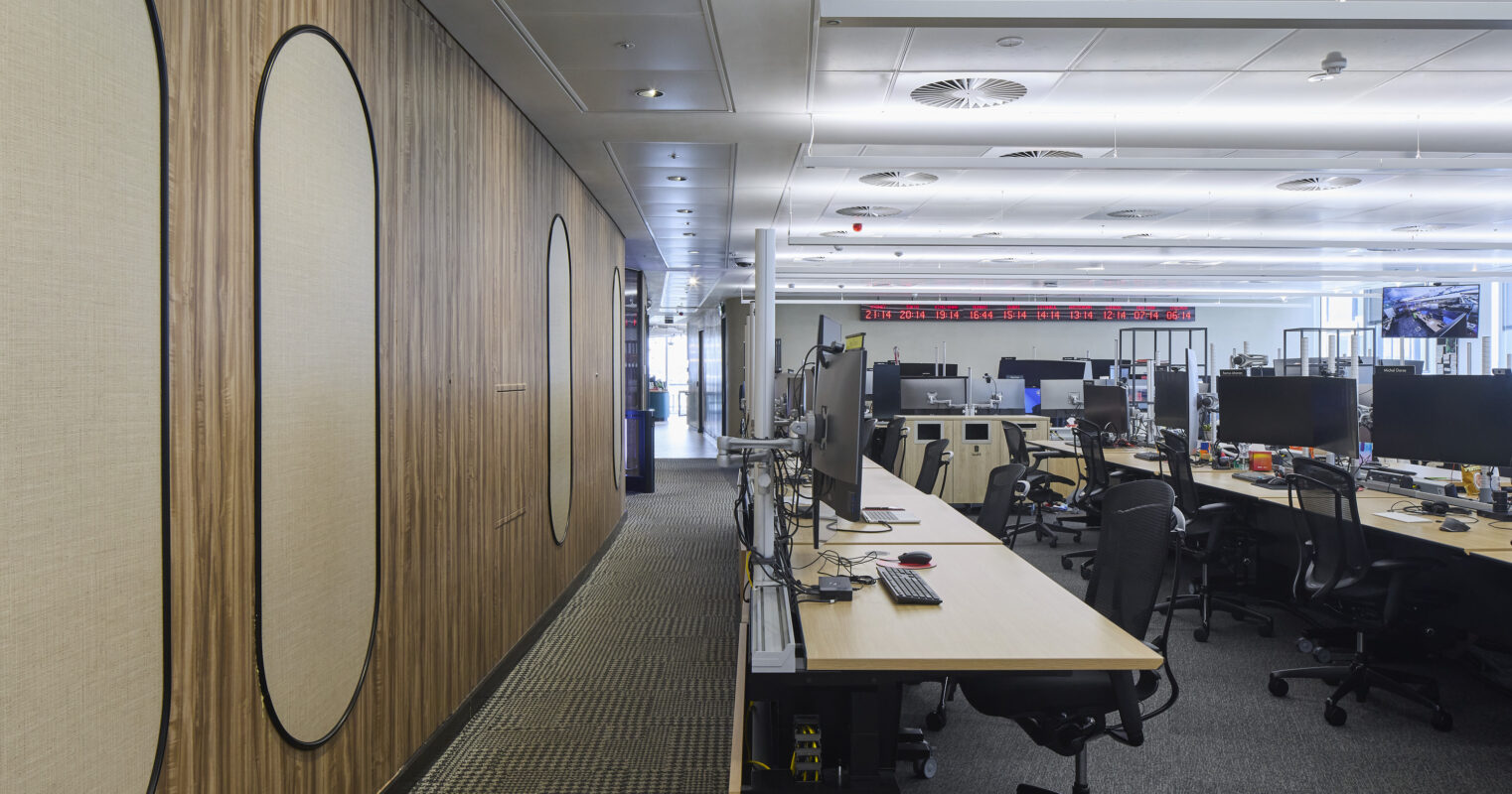 Modern office interior featuring sleek wooden wall panels with oval insets, creating a texture-rich backdrop. A row of minimalist workstations with ergonomic chairs and multiple monitors provide a functional workspace. The open layout enhances collaboration, while strategic lighting offers a bright, inviting atmosphere.