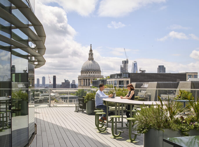 A vibrant rooftop terrace features modern seating arrangements surrounded by lush greenery. Two individuals engage in conversation at a sleek table, with a backdrop showcasing London’s iconic St. Paul's Cathedral and contemporary skyscrapers, blending urban life with outdoor relaxation in a stylish setting.