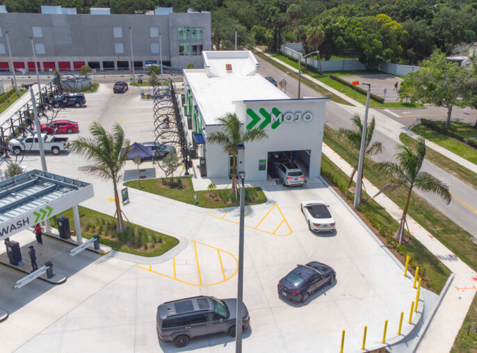 A modern car wash facility features a minimalist architectural design with a bright white exterior and bold green branding. The layout includes several car washing stations, palm trees for landscaping, and designated parking areas. Enhanced with efficient elements, the design prioritizes both functionality and aesthetic appeal.