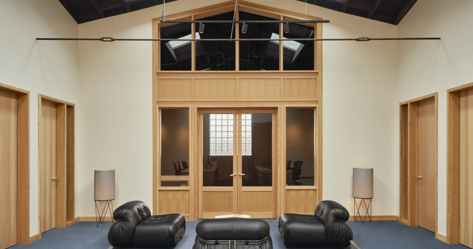 Spacious interior featuring a high, wood-paneled ceiling with skylights. The room includes minimalist black lounge furniture, providing a modern contrast to the warm wooden accents. Framed glass doors lead to adjacent spaces, while sleek lamps enhance the contemporary aesthetic. Neutral tones create an inviting atmosphere.