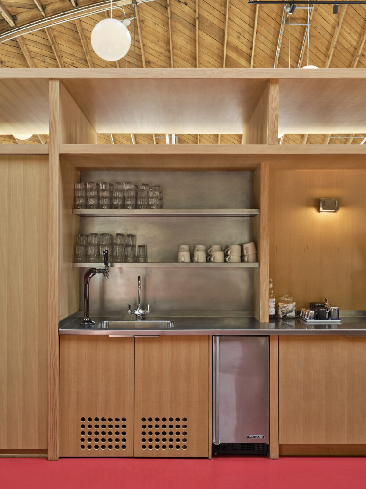 Warm wooden cabinetry complements sleek stainless steel shelving in this modern kitchen space. The open layout features minimalist design elements, with glassware and dishware neatly arranged. A bright red floor adds a bold contrast to the natural materials, enhancing the overall aesthetic of functionality and simplicity.