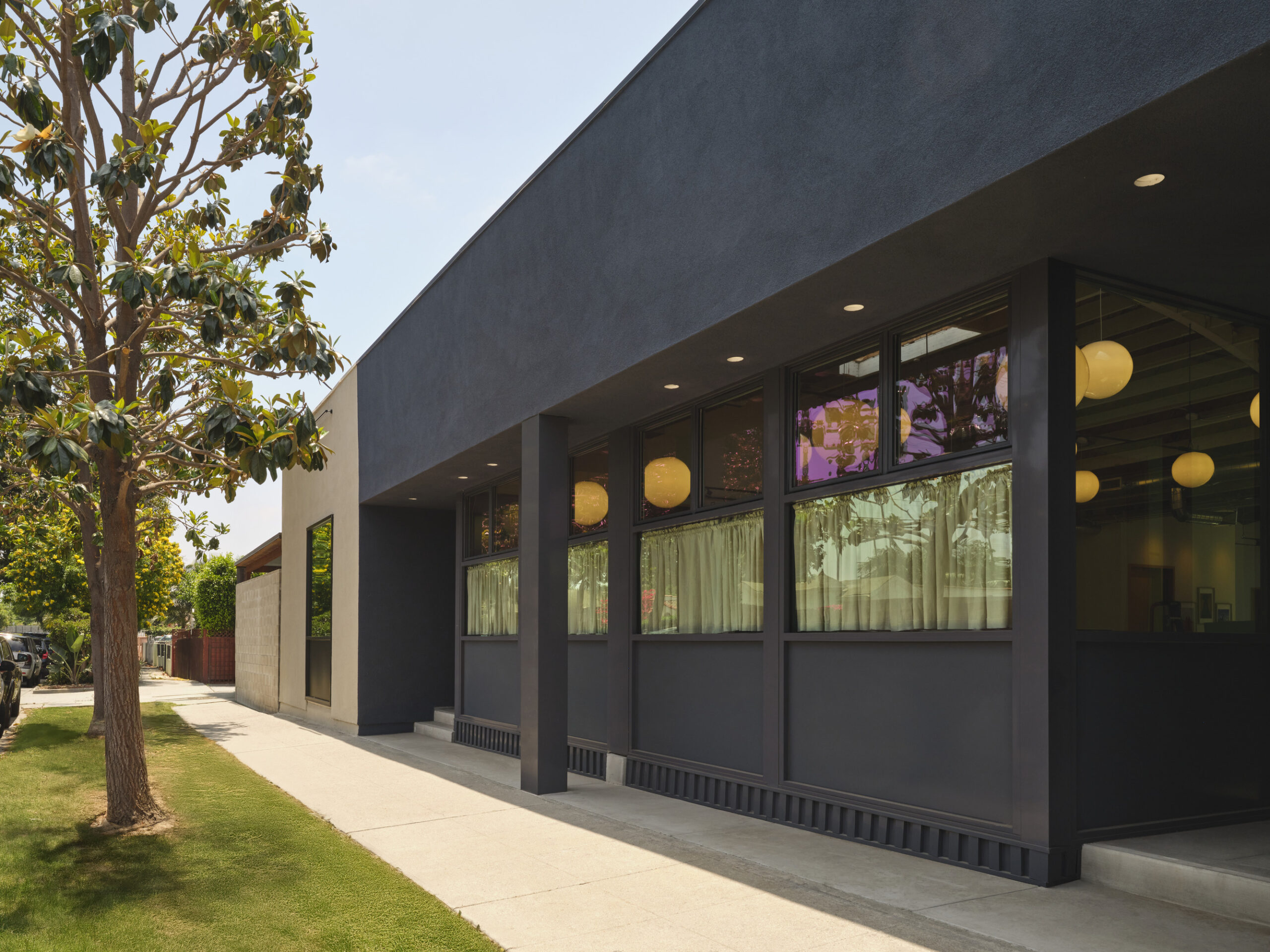 A modern architectural facade featuring a sleek, dark exterior with large windows, allowing natural light to permeate the interior. The design incorporates minimalist features, complemented by surrounding greenery. Soft pendant lighting enhances the inviting ambiance, establishing a harmonious connection between the structure and its environment.