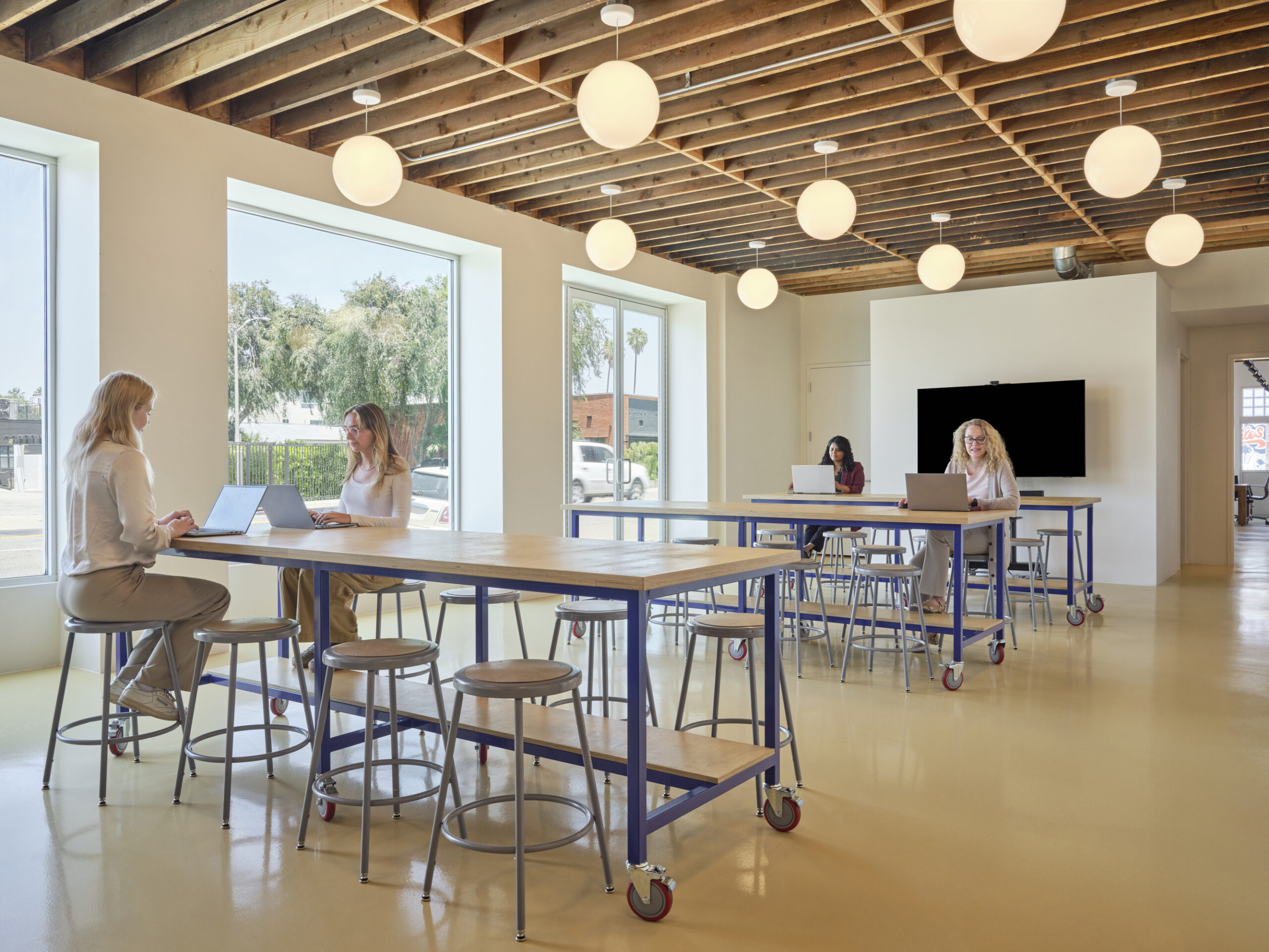 Bright, collaborative workspace featuring an open layout with large windows allowing natural light to flood in. Modern wooden beams contrast with sleek, minimalist furniture. High-top tables with metal frames invite teamwork, while round pendant lights add a warm ambiance, enhancing the creativity and productivity of the environment.