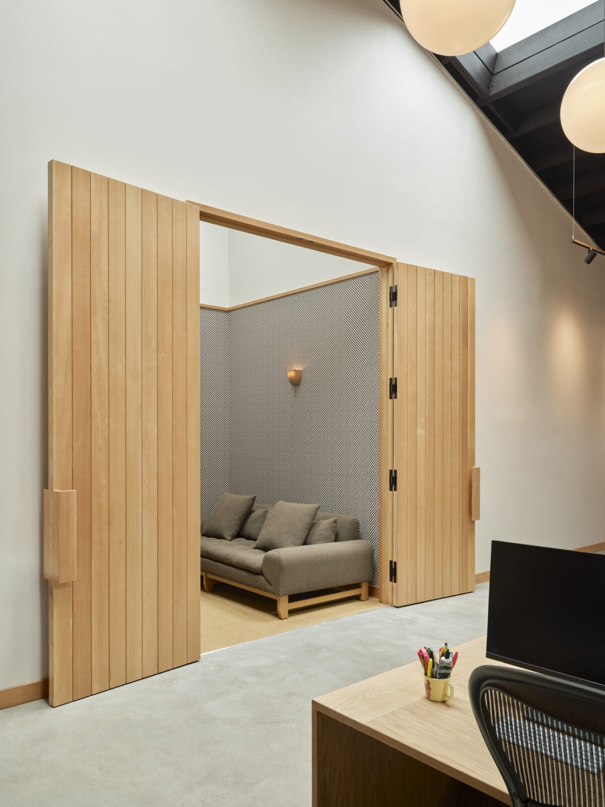 Warm wooden bi-fold doors open to a cozy lounge area, featuring a textured gray wallpaper and soft, modern seating. The soft lighting complements the minimalist aesthetic, while the adjacent workspace showcases a functional desk, emphasizing the balance between professional and inviting interior spaces.