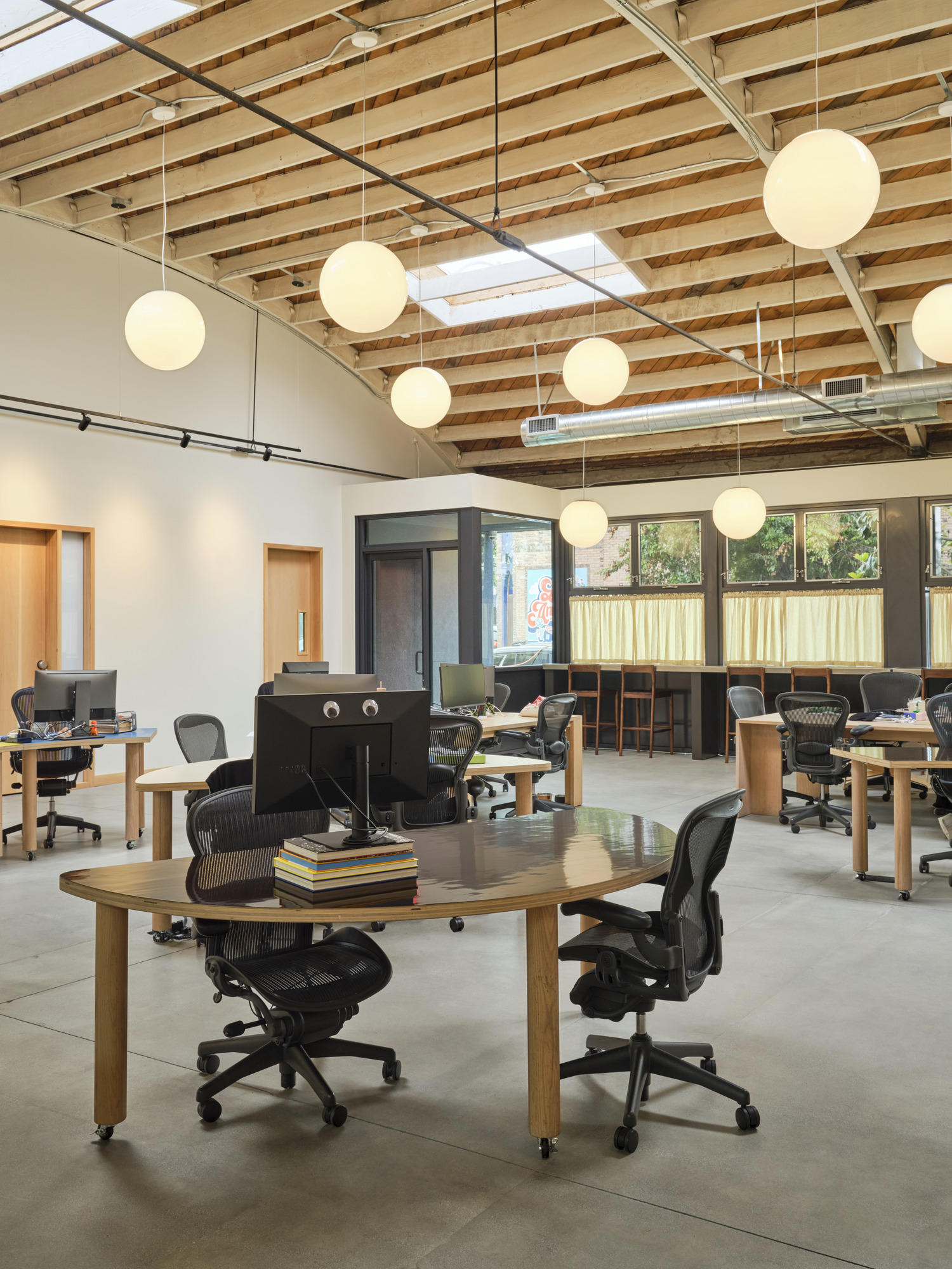 Open-plan office featuring modern workstations with ergonomic chairs, set against a backdrop of exposed wooden beams and a high ceiling. Large round pendant lights provide ambient lighting, while natural light flows through skylights and large windows, creating a bright and inviting work environment.