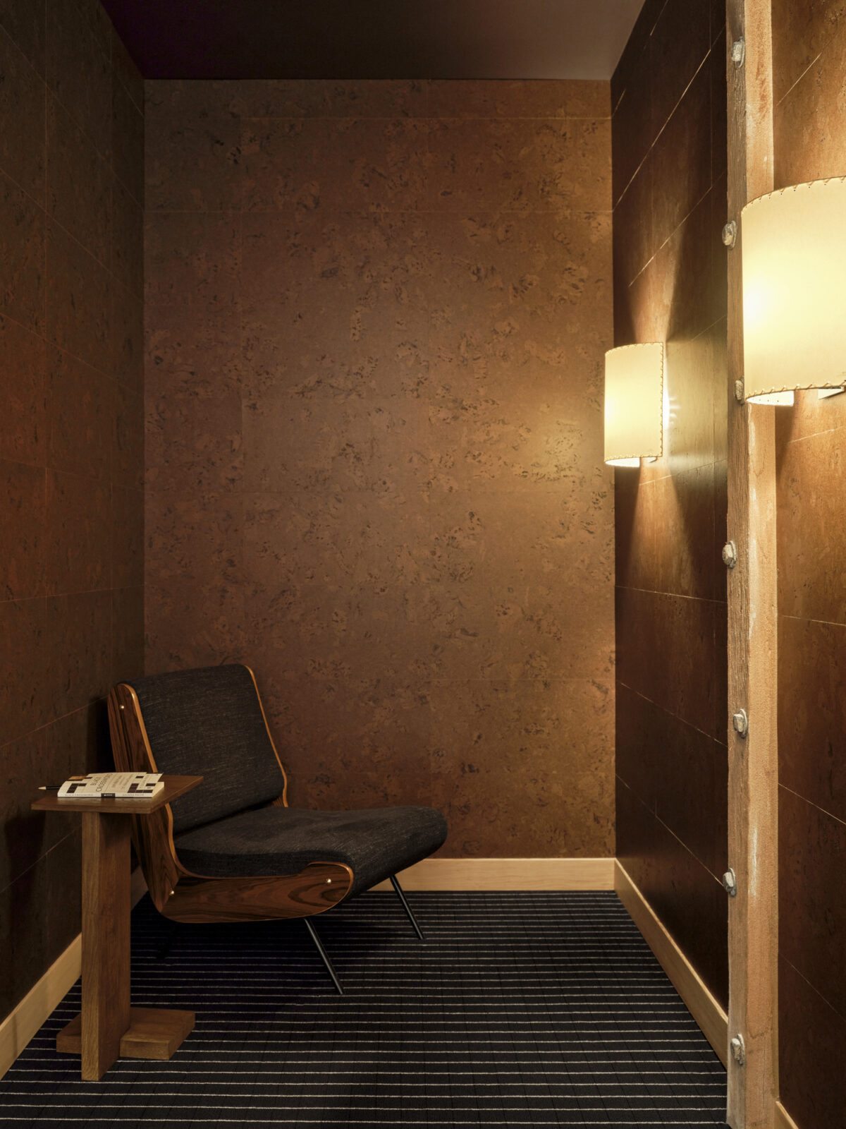 A cozy corner featuring a mid-century modern lounge chair in dark fabric, accompanied by a minimalist wooden side table. The walls are clad in rich, textured cork, creating a warm atmosphere. Soft wall sconces illuminate the space, highlighting the contrast between the dark finishes and the striped flooring.