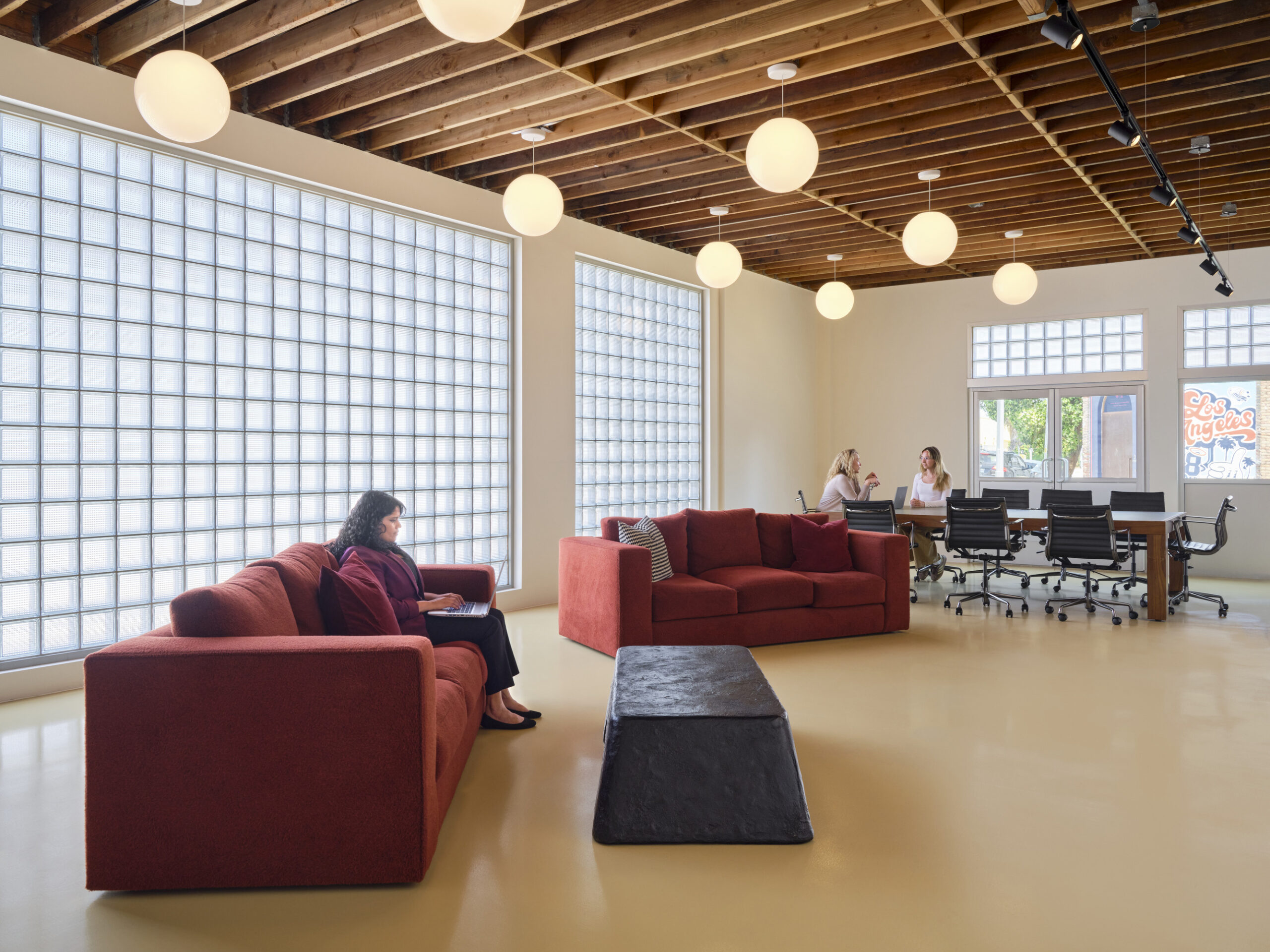 Spacious office interior featuring a modern design with exposed wooden beams and ample natural light. Two lounging areas, including a vibrant red sofa, encourage collaboration. Glass block windows enhance privacy while allowing light. In the background, a meeting area with a large table and ergonomic chairs promotes productivity.