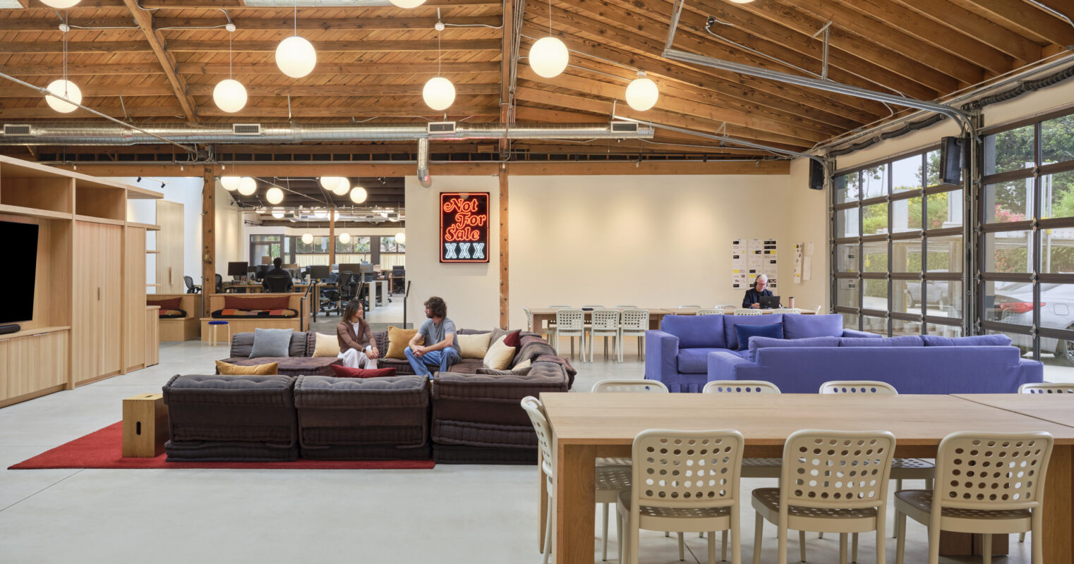 A modern, spacious interior featuring high, exposed wooden ceilings and ample natural light. Cozy seating areas with a mix of sofas and dining tables invite collaboration. The neutral color palette contrasts with vibrant furniture, while unique pendant lights enhance the open atmosphere, creating a welcoming workspace.