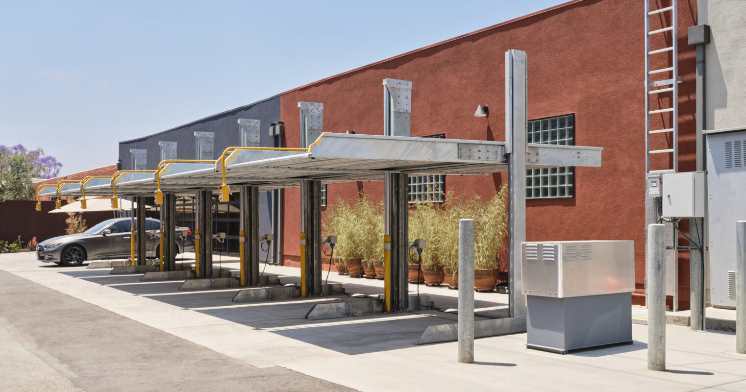 A contemporary carport design featuring sleek metal structures, providing shade and protection for vehicles. The background showcases a vibrant facade with red and blue walls, punctuated by large windows. The space is neatly organized with industrial elements and lush greenery, blending functionality with modern aesthetics.