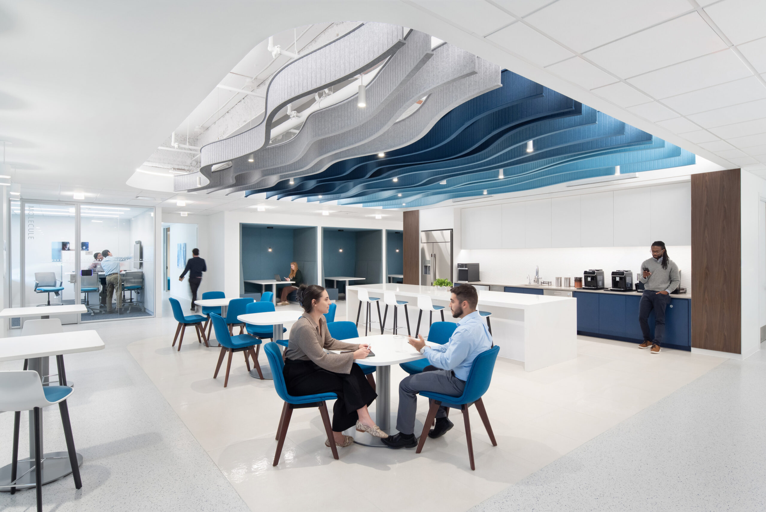 A modern office interior featuring a collaborative space with sleek, blue accent furniture. Innovative wave-like ceiling panels create visual interest, while a multifunctional kitchenette serves as a focal point. Natural light enhances the open layout, encouraging teamwork and casual meetings among employees in stylish surroundings.