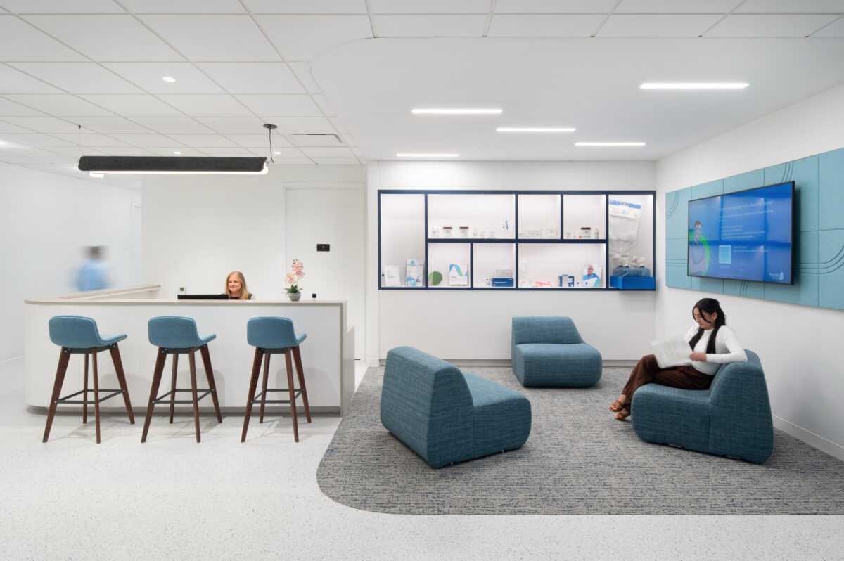 Bright, modern reception area featuring curved blue seating arranged on a textured gray carpet. A sleek white reception desk with backlighting and a digital display on the wall showcases the brand's identity. Natural light enhances the inviting atmosphere, encouraging a comfortable waiting experience for guests.