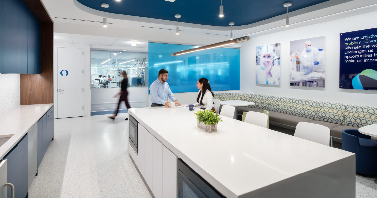 Bright, modern corporate kitchen and lounge area featuring sleek white quartz countertops, wooden cabinetry, and decorative tile walls. A long communal table encourages collaboration, while pendant lighting highlights the space. Blue accents create a fresh atmosphere, complemented by wall-mounted screens displaying inspirational visuals. A casual yet professional design fosters creative engagement.