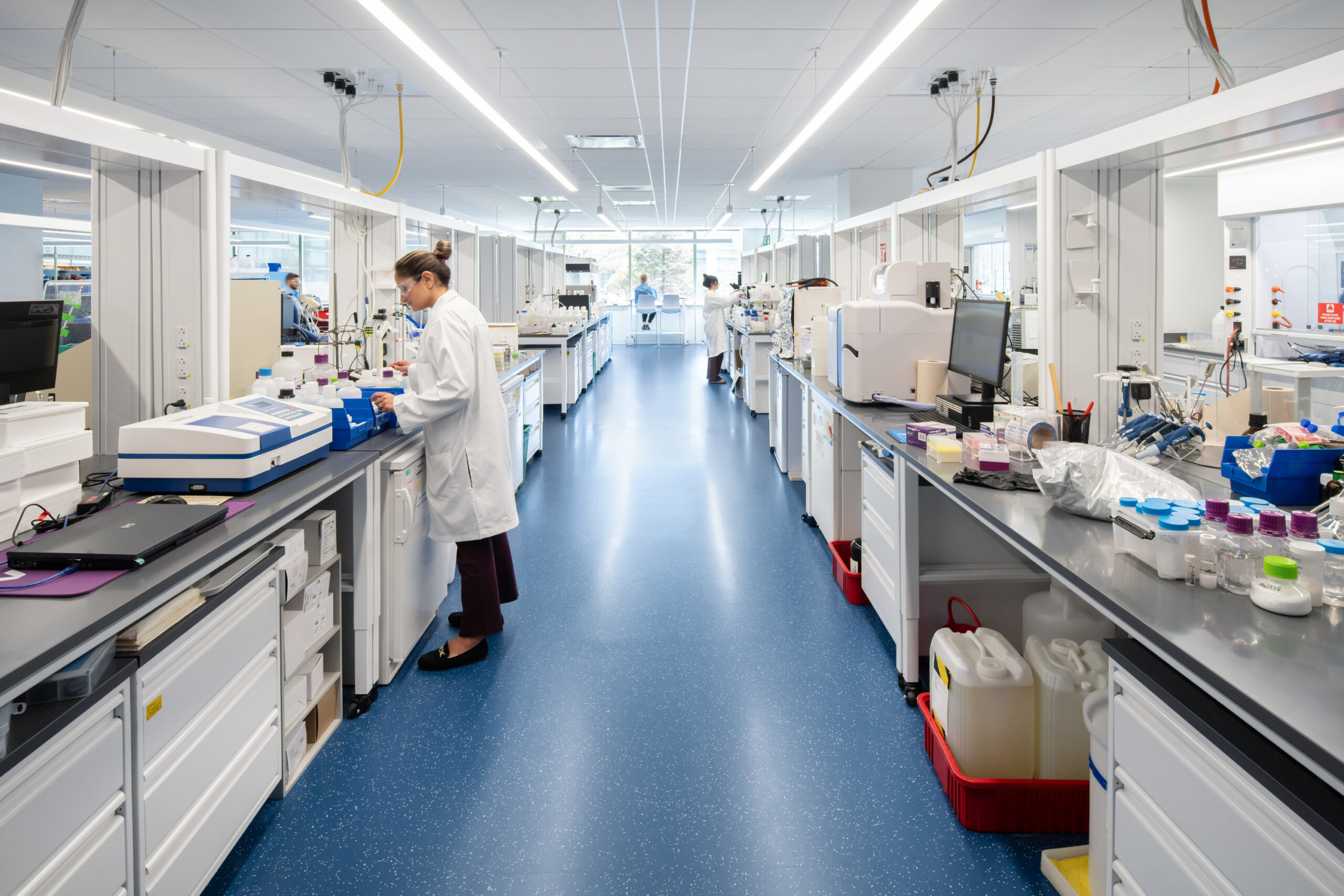 A modern laboratory featuring spacious workstations equipped with advanced scientific instruments, including centrifuges and spectrophotometers. Natural light floods the area through large windows, enhancing the clean, minimalist design while promoting a collaborative atmosphere among researchers in lab coats. Bright blue flooring contrasts with white cabinetry, ensuring a functional yet visually appealing environment.