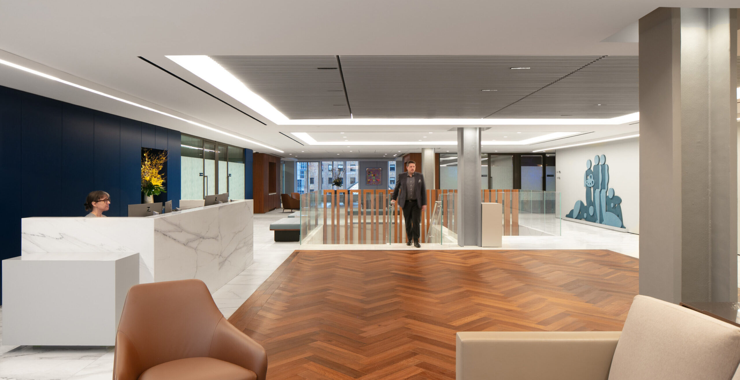 Modern office lobby showcasing a blend of elegance and functionality. Rich herringbone wood flooring contrasts with smooth marble surfaces, while a dark blue accent wall adds depth. A sleek reception desk complements contemporary furnishings, and strategically placed art enhances the open space, fostering a welcoming atmosphere for visitors.