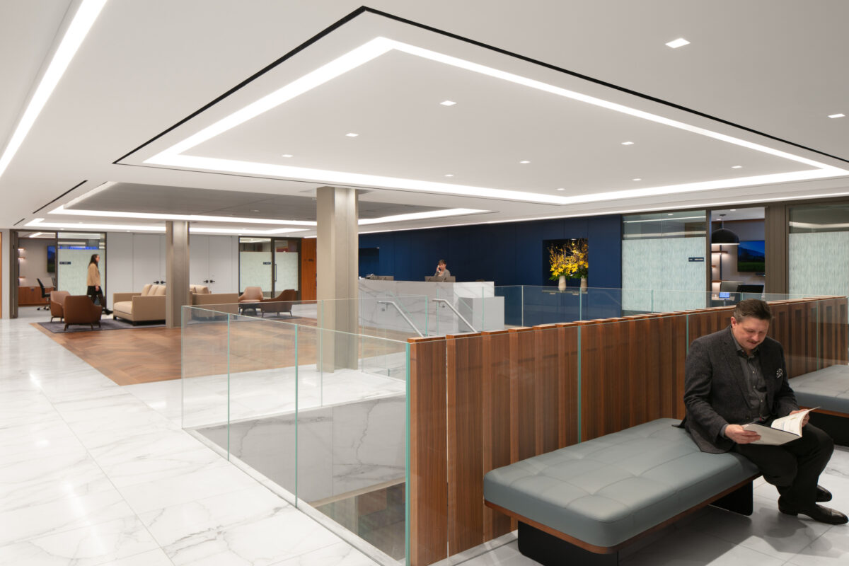 A modern reception area showcasing sleek design elements, featuring a marble floor and warm wood accents. Soft seating spaces are integrated into the layout, with a glass partition providing openness. Illumination highlights architectural features, while a blue accent wall adds a contemporary touch, creating an inviting atmosphere.