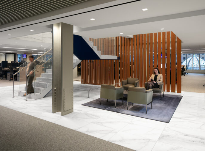 Sophisticated office lobby featuring a sleek, modern staircase with a glass railing and wooden slat partitions. Plush seating areas in neutral tones offer comfort, complemented by a stylish area rug. Large windows allow natural light to enhance the open layout, creating a welcoming and collaborative atmosphere.