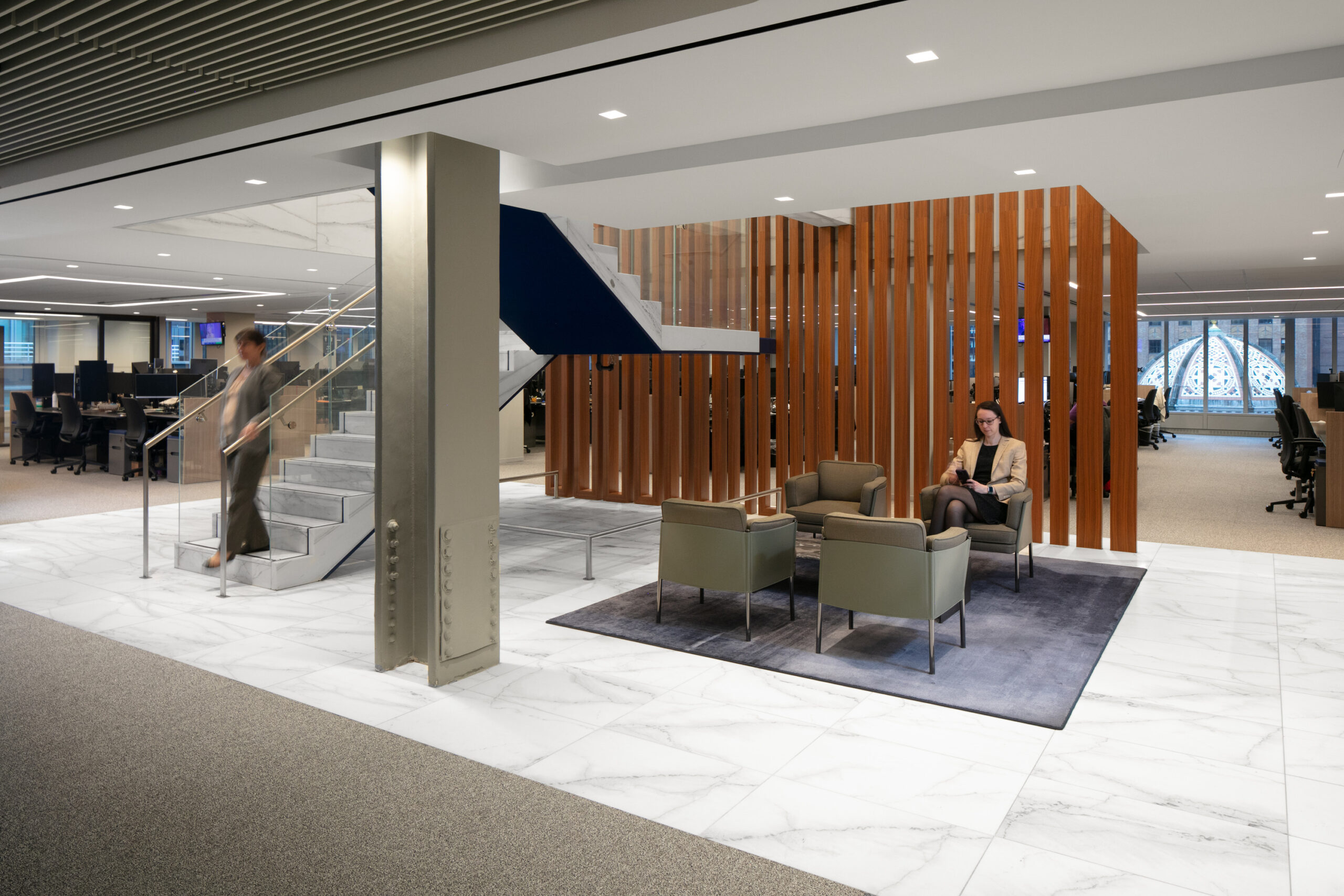 Sophisticated office lobby featuring a sleek, modern staircase with a glass railing and wooden slat partitions. Plush seating areas in neutral tones offer comfort, complemented by a stylish area rug. Large windows allow natural light to enhance the open layout, creating a welcoming and collaborative atmosphere.