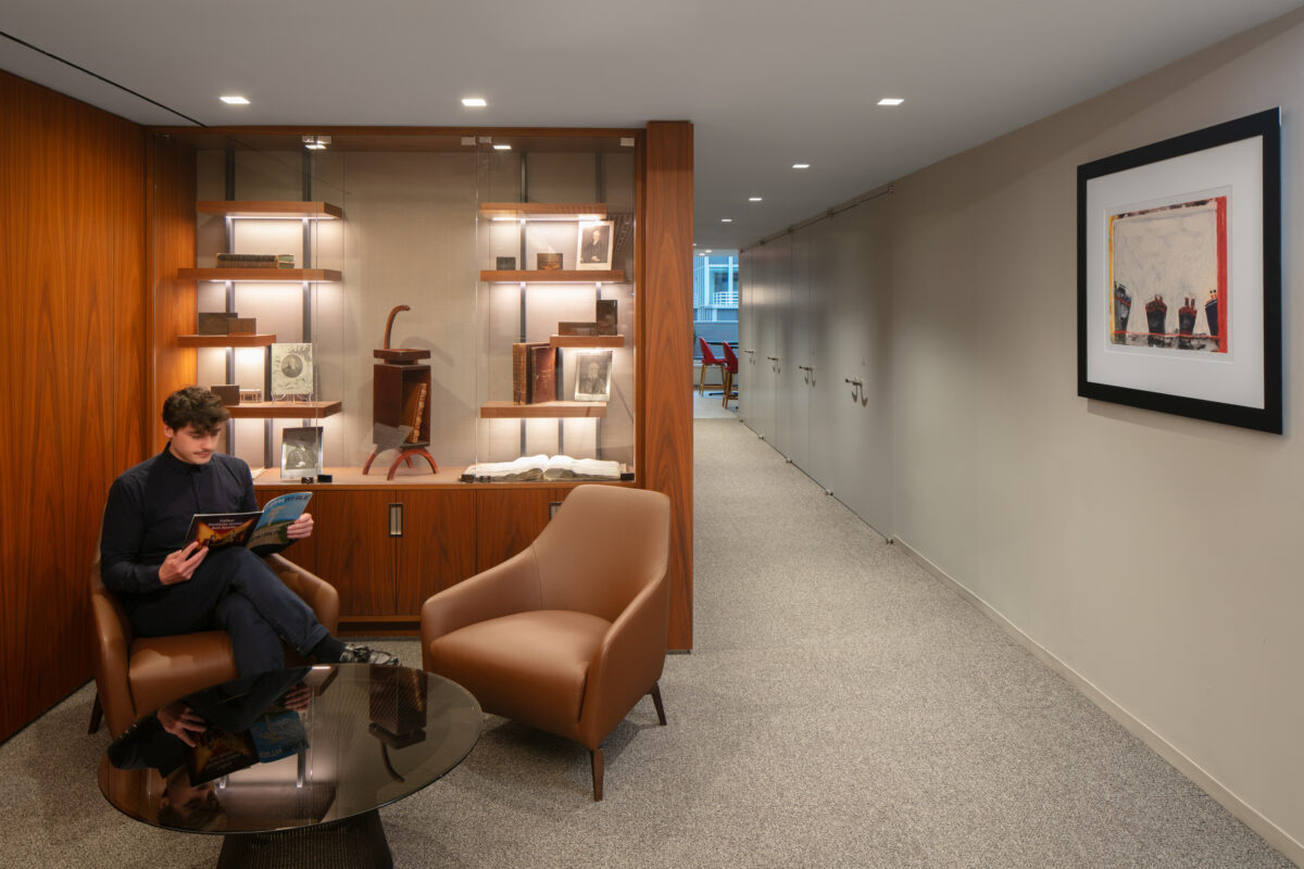 A stylish reading nook features a comfortable brown armchair next to a chic circular black table. On a sleek wooden wall, illuminated shelves display vintage books and art pieces. A relaxed individual engages with a magazine, embodying a blend of modern comfort and timeless elegance in this thoughtfully designed interior.