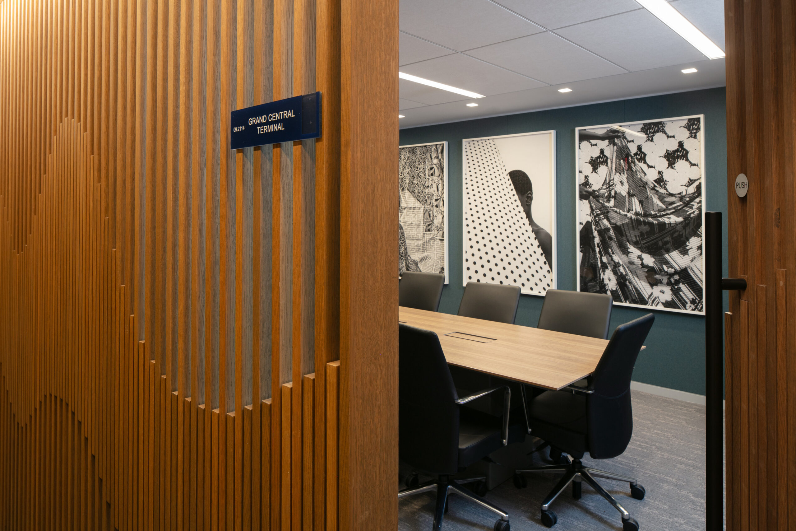 Modern conference room featuring a long wooden table surrounded by ergonomic black chairs. The backdrop showcases bold, framed artworks with abstract patterns. Textured wooden slats on the entrance door add warmth and sophistication, creating a welcoming atmosphere that combines functionality with contemporary design.