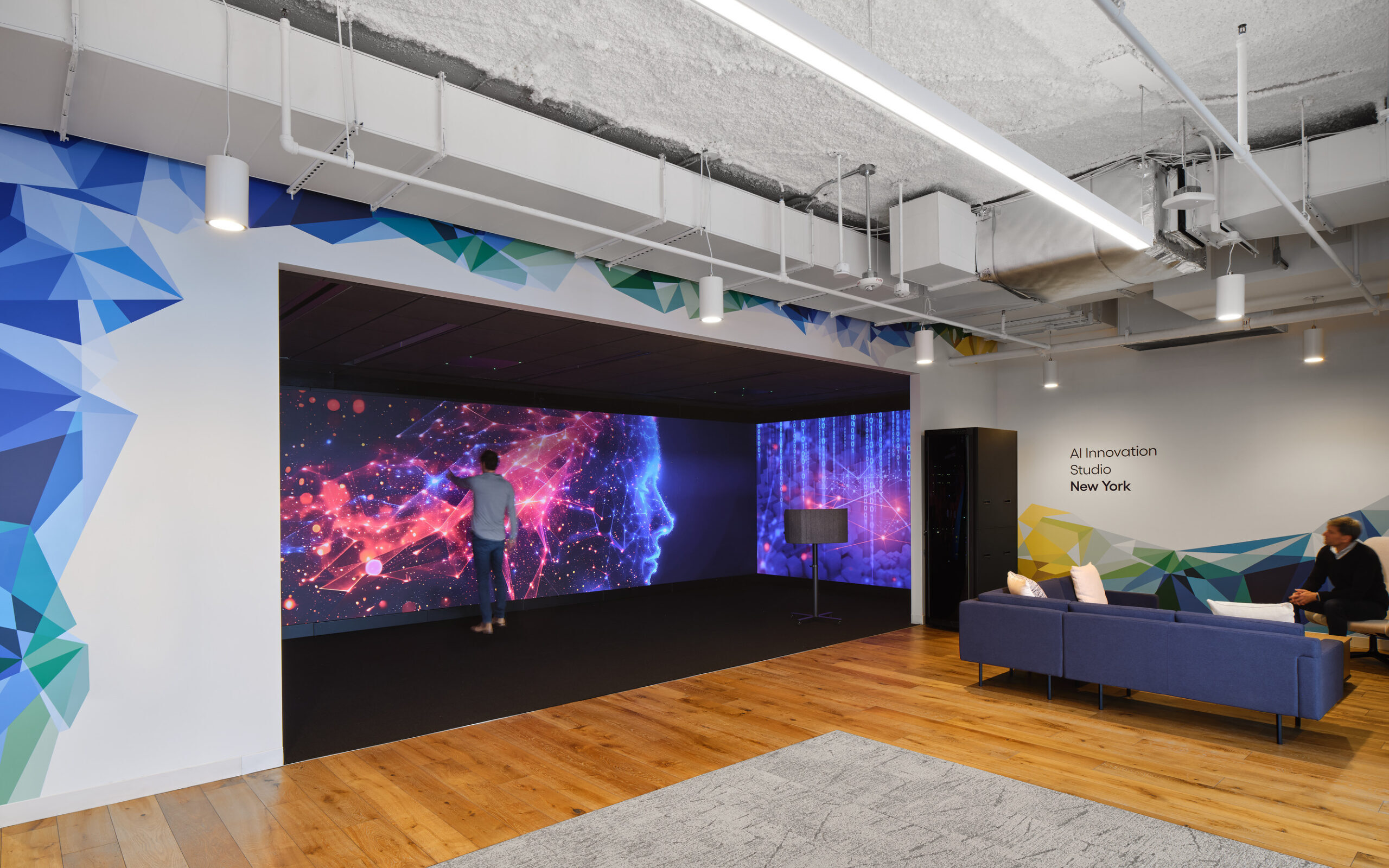 A modern innovation studio featuring dynamic geometric wall graphics in vibrant colors. The space includes a large projection area displaying abstract visuals, fostering creativity and collaboration. Two individuals engage with the technology and ambiance, while contemporary furniture enhances the inviting atmosphere with wooden flooring and textured ceiling elements.