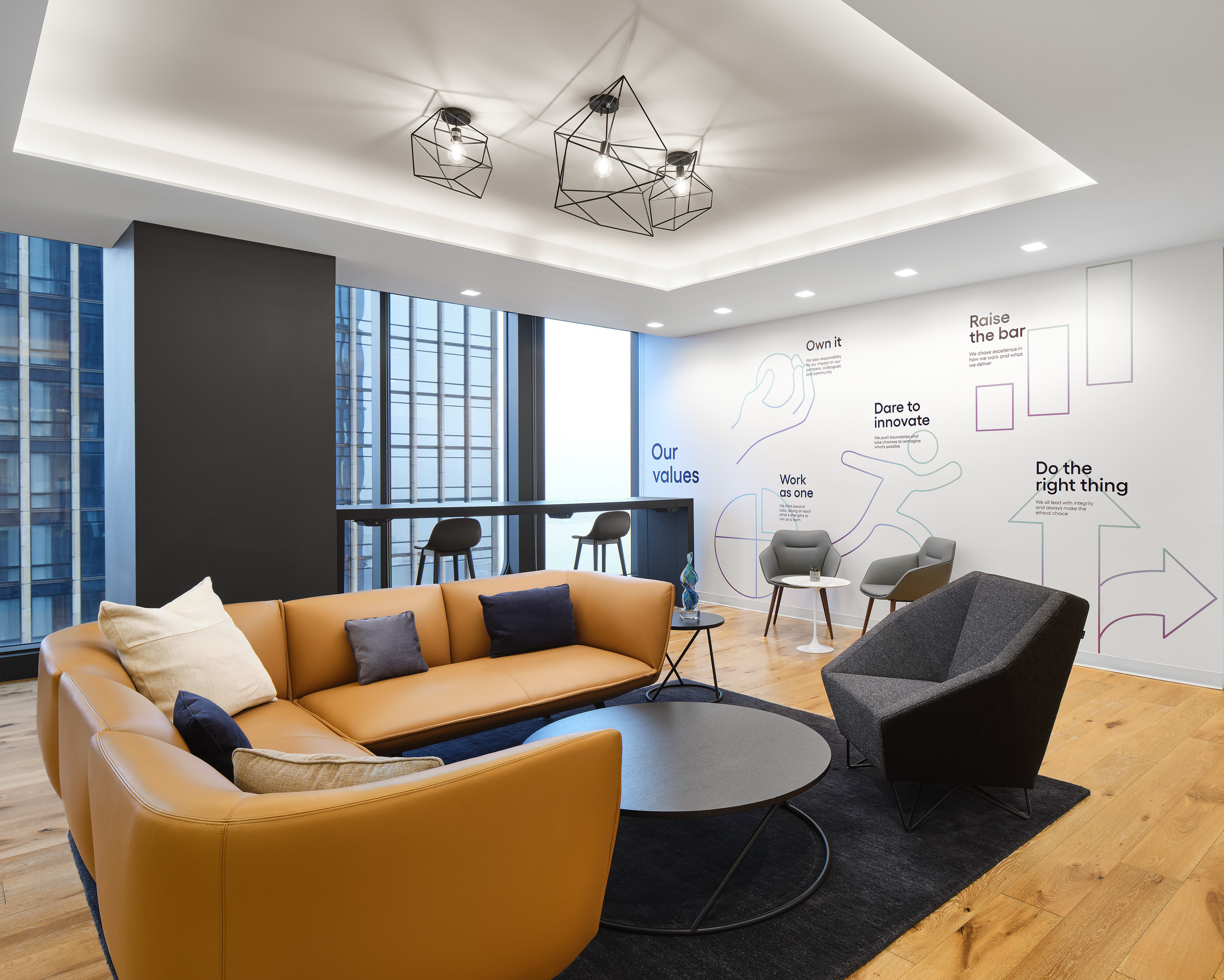 A modern office lounge features a sleek, curvilinear orange sofa paired with dark blue pillows, complemented by a geometric black armchair. A round black coffee table sits atop a textured rug, while floor-to-ceiling windows provide ample natural light. Inspirational wall graphics highlight company values and promote a collaborative workspace.