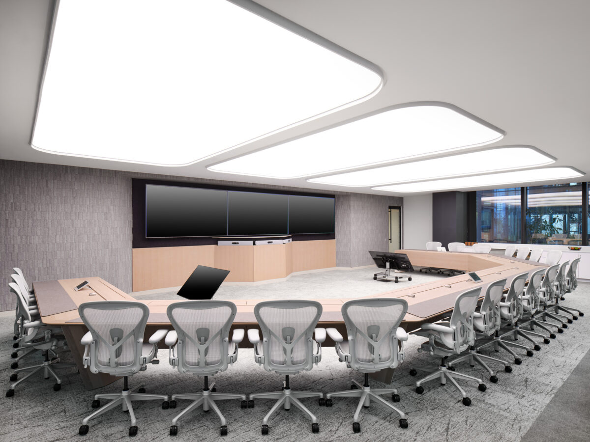 A modern conference room featuring a large elliptical wooden table surrounded by ergonomic office chairs. Sleek overhead lighting creates an inviting ambiance, complemented by three large screens for presentations. The neutral color palette and large windows promote a collaborative and professional environment, ideal for meetings and discussions.