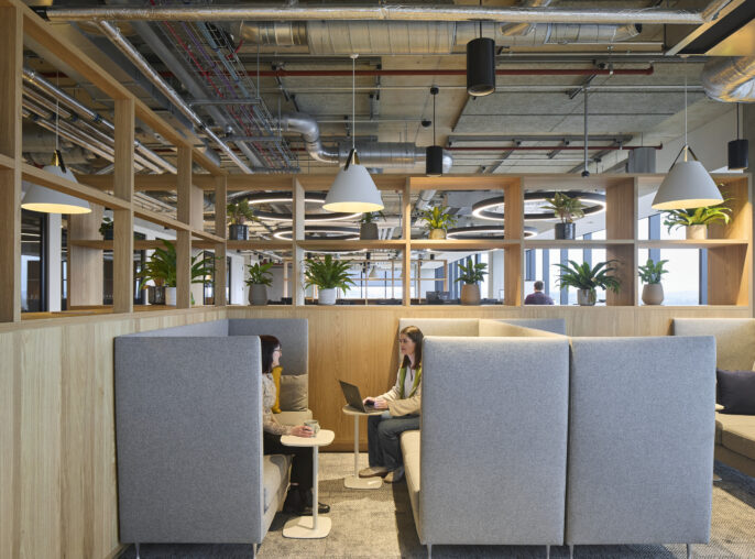 Contemporary office space featuring modular seating with high backrests, promoting privacy. Warm wooden panels and lush greenery enhance the inviting atmosphere. Industrial-style elements, including exposed ductwork and pendant lighting, contrast with the soft furnishings. Workstations are designed for collaboration and comfort, fostering productivity and creativity.