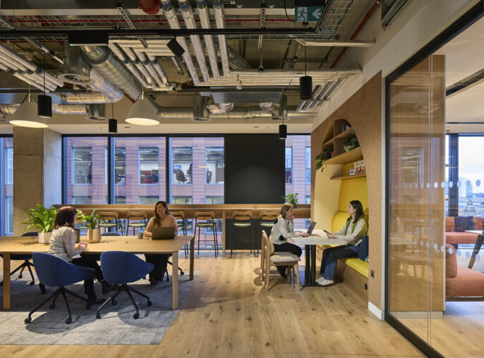 An open-plan office space features a blend of collaborative and casual seating areas. Modern wooden furniture complements the warm tones of natural light from large windows, while decorative plants enhance the inviting atmosphere. Distinctive arch-shaped shelving adds visual interest, emphasizing functional design.
