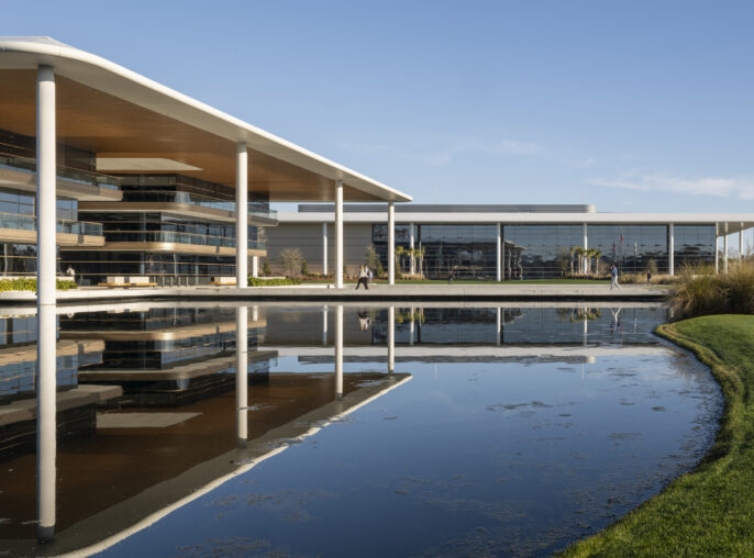 Modern corporate campus featuring expansive glass façades, sweeping rooflines, and white columns overlooking a reflective pond, with landscaped grounds and pedestrian walkways under a clear blue sky.