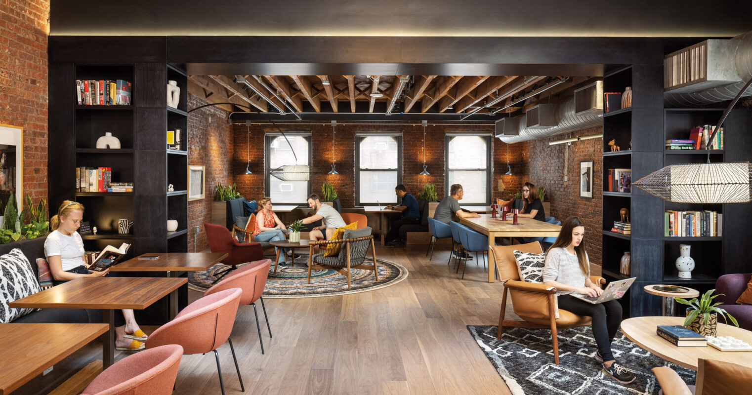A modern, cozy co-working space bustling with professionals engaged in various activities, from working on laptops to reading, in a room with exposed brick walls, stylish furnishings, and warm lighting.