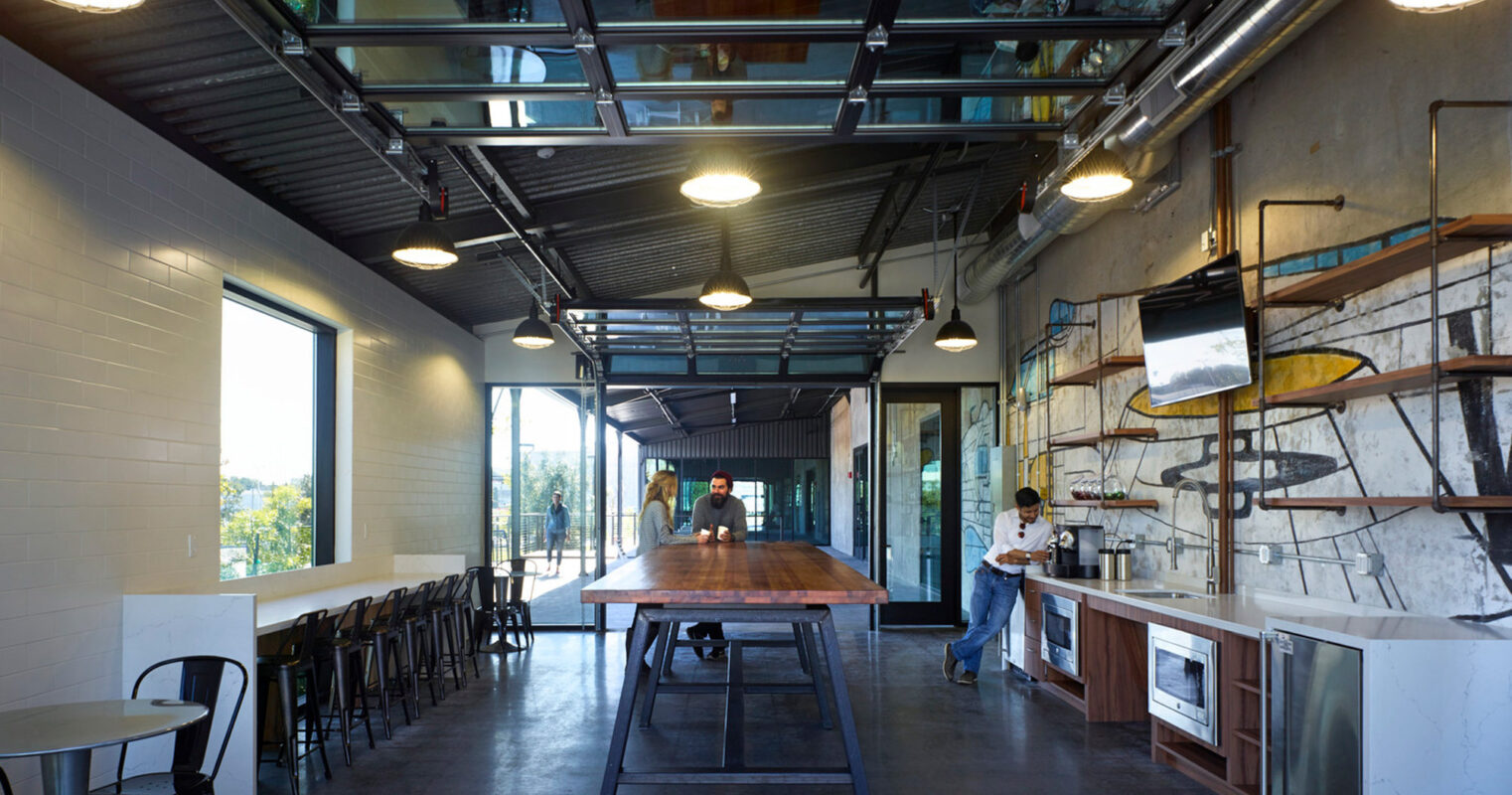 Modern industrial-themed café interior featuring exposed ceiling pipes, track lighting, and a polished concrete floor. Large windows and a glass ceiling section flood the space with natural light. A communal wooden table, bar stools, and counter service area emphasize a casual, social atmosphere.