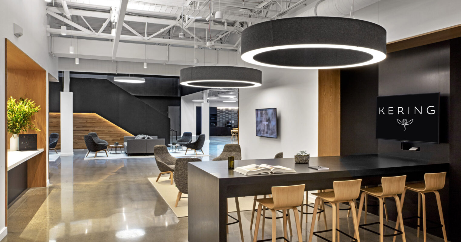 Modern office lobby with a monochromatic color scheme, featuring circular overhead lighting and a black reception desk with a branded logo. Architectural elements include exposed ceiling beams and a wooden staircase leading to an upper level. Contemporary lounge seating is strategically placed for casual encounters.