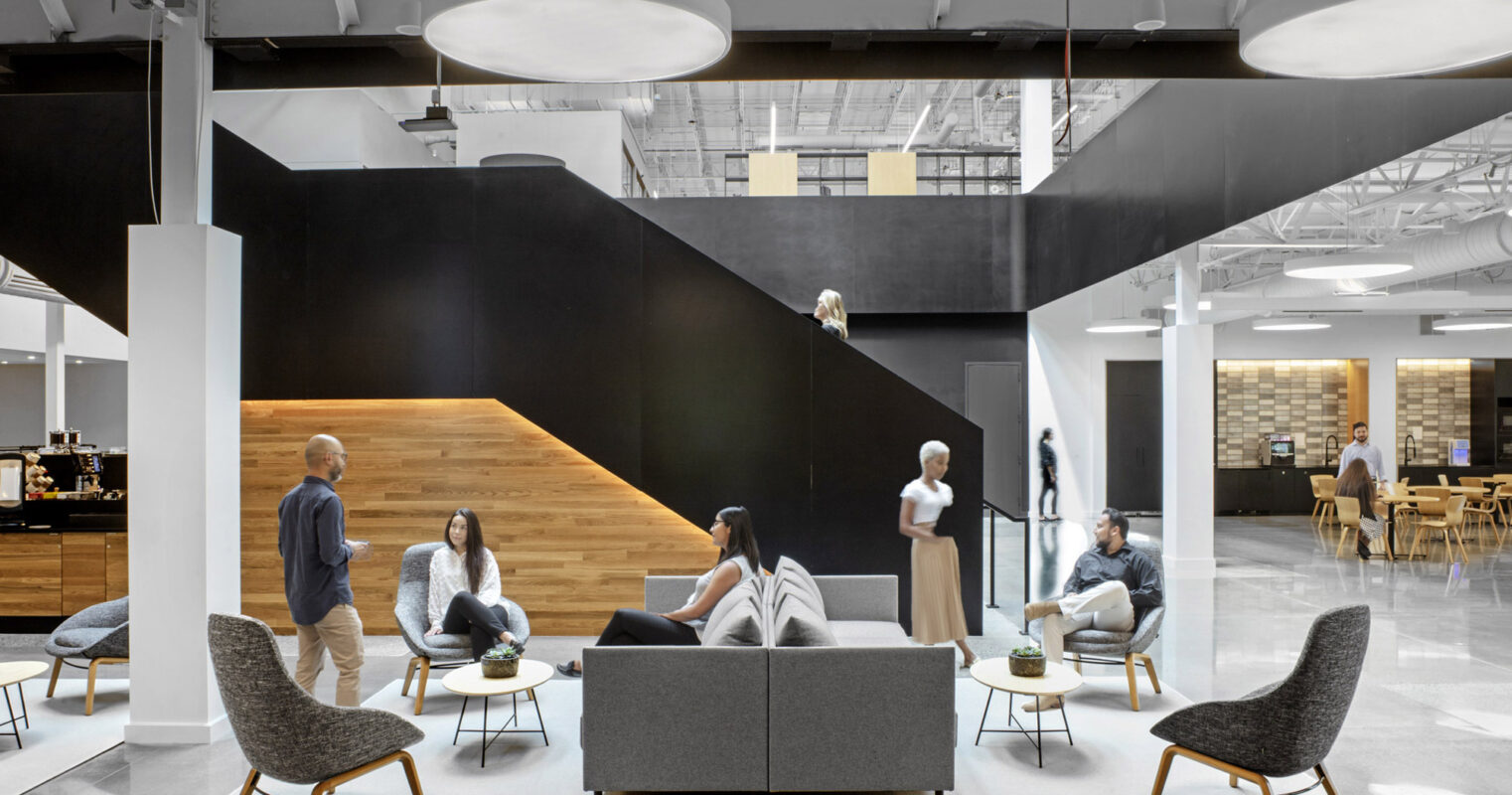 Modern office lounge with monochromatic color scheme, featuring a striking black staircase centerpiece and eclectic seating arrangements with Scandinavian-inspired chairs, promoting collaborative and individual relaxation spaces.