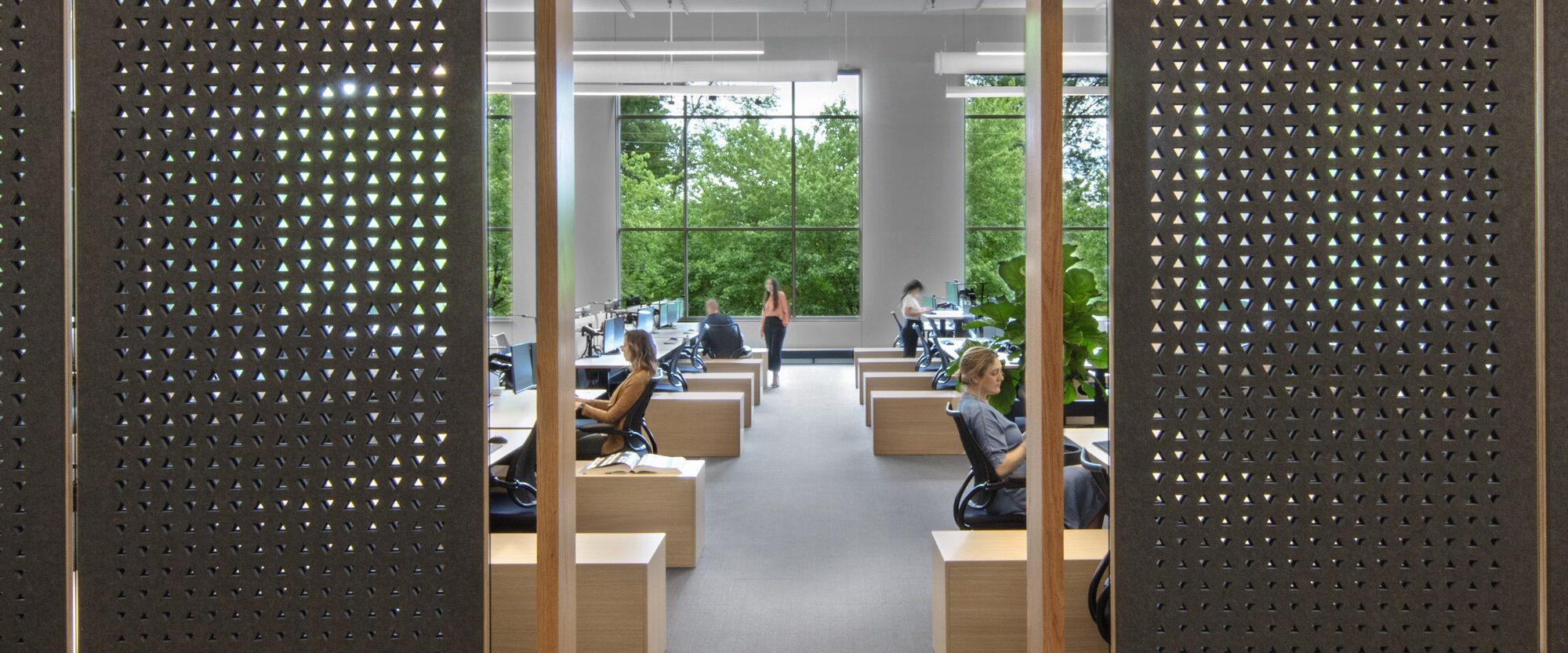 Modern open-plan office space featuring wooden privacy screens with intricate geometric cutouts, ergonomic workstations, and floor-to-ceiling windows providing ample natural light.