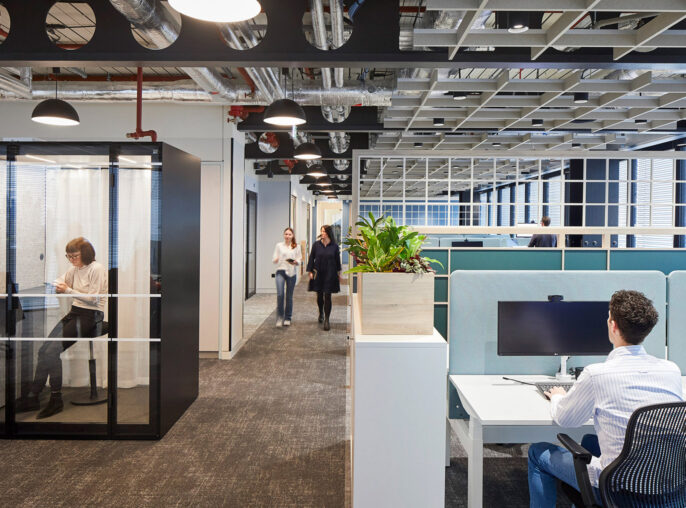 Modern office interior featuring an open floor plan with individual workstations, glass-walled meeting rooms, exposed ceiling beams, and vibrant blue privacy booths. Natural light and pedestrian movement evoke a dynamic work environment.